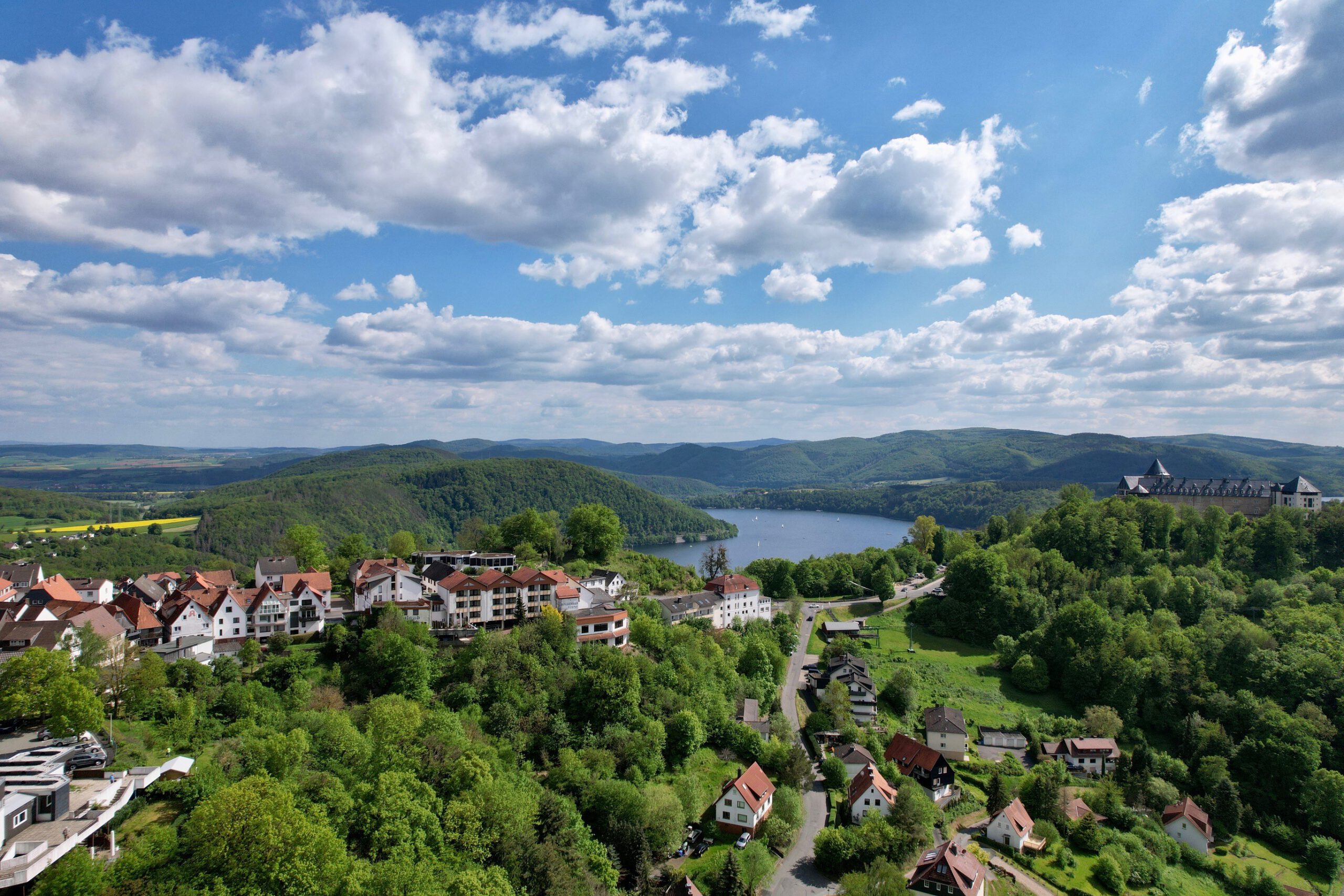 Blick von oben auf Waldeck, im Hintergrund der Edersee, davor auf einem Berg das Schloss Waldeck, blauer Himmel mit Wolken