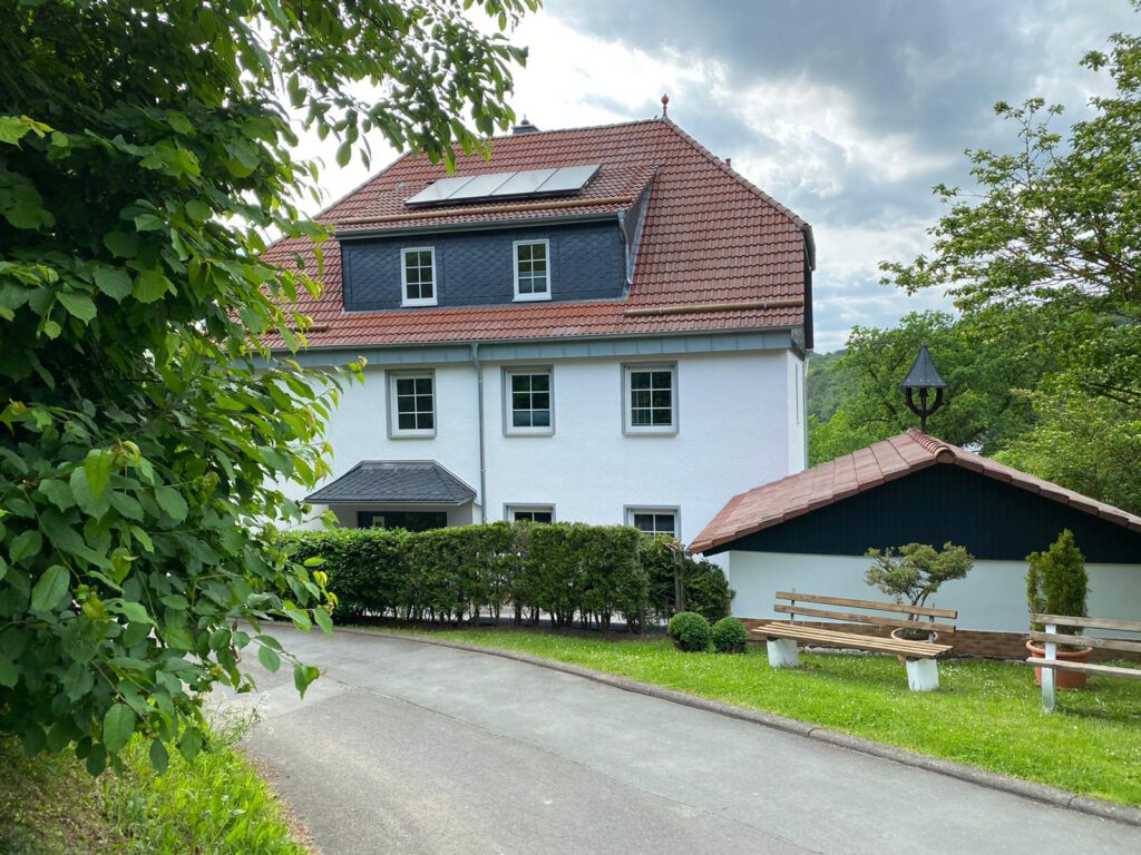 Ansicht vom Bergschlösschen; Im unteren Bild eine kleine Straße, die zum Haus führt; vor dem Haus eine grüne Hecke mit zwei Bänken